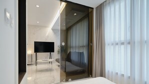 A modern hotel room with a glass partition, a flat-screen TV, a round table, and a bed with a view through sheer curtains. - UH FLAT THE SEOCHO (Seoul)