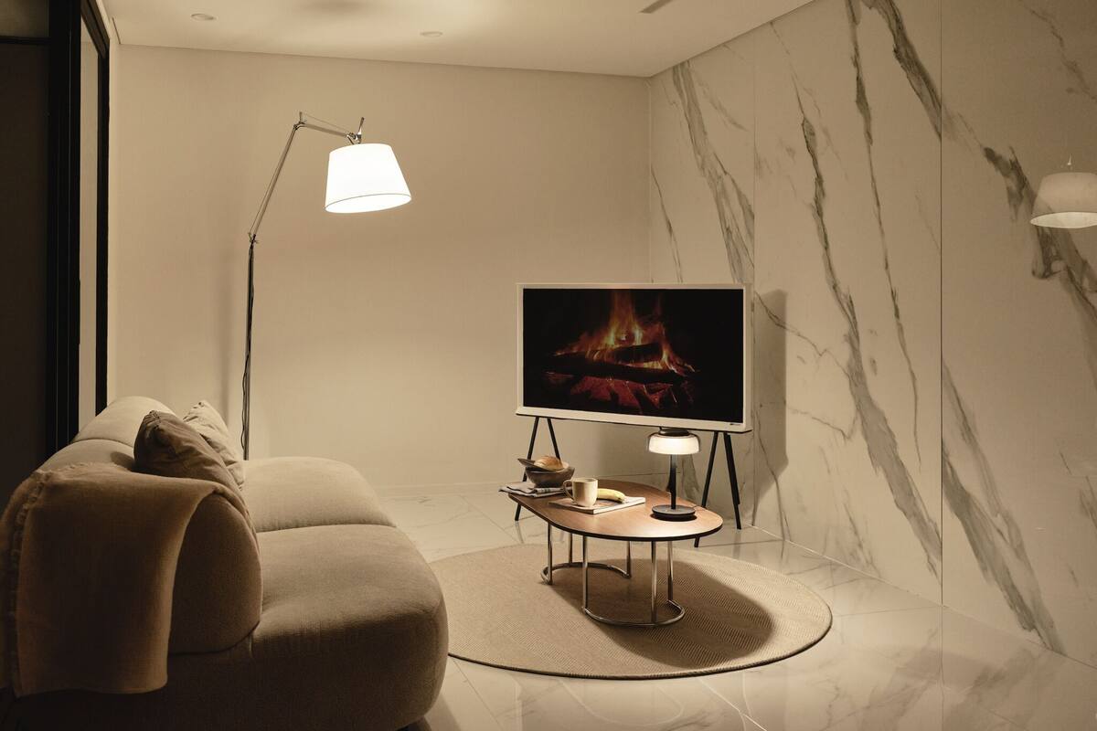 A modern living room with a sofa, a television displaying a fire, a floor lamp, and a marble wall.
