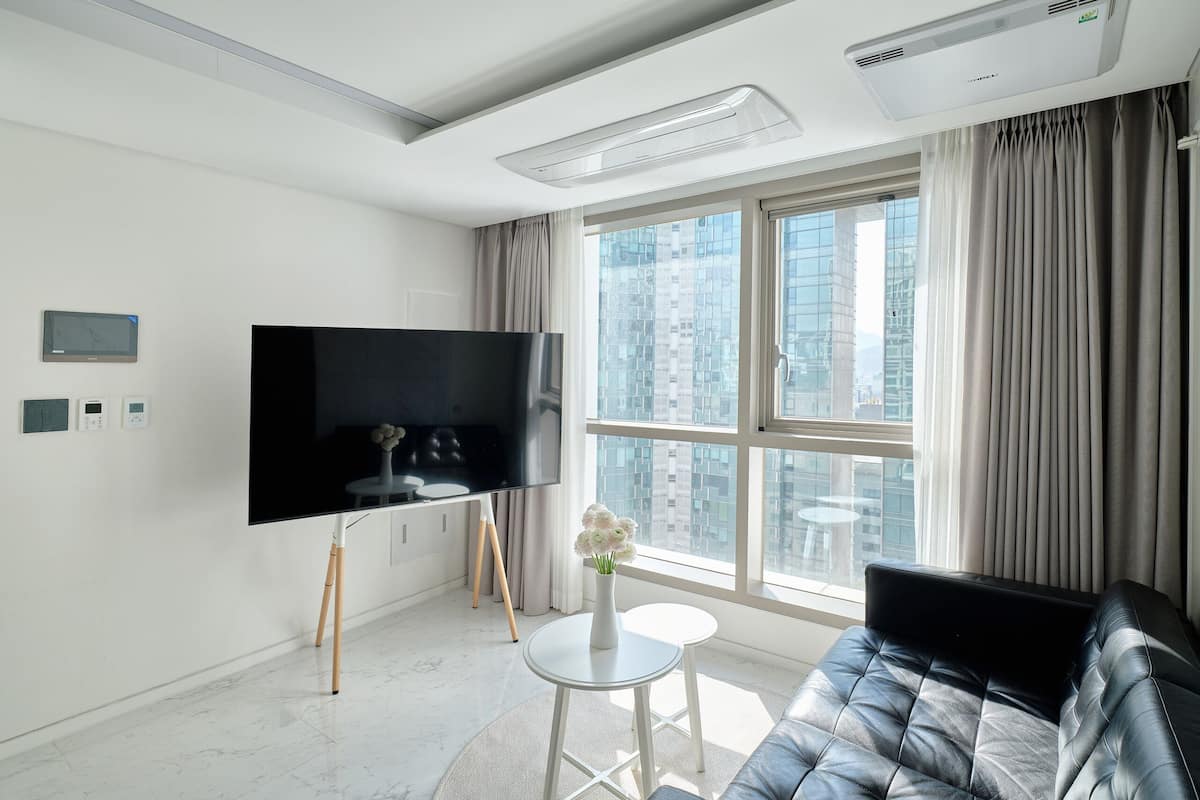 A modern living room with a flat-screen TV, a black leather sofa, a small white table, and a window with a city view.