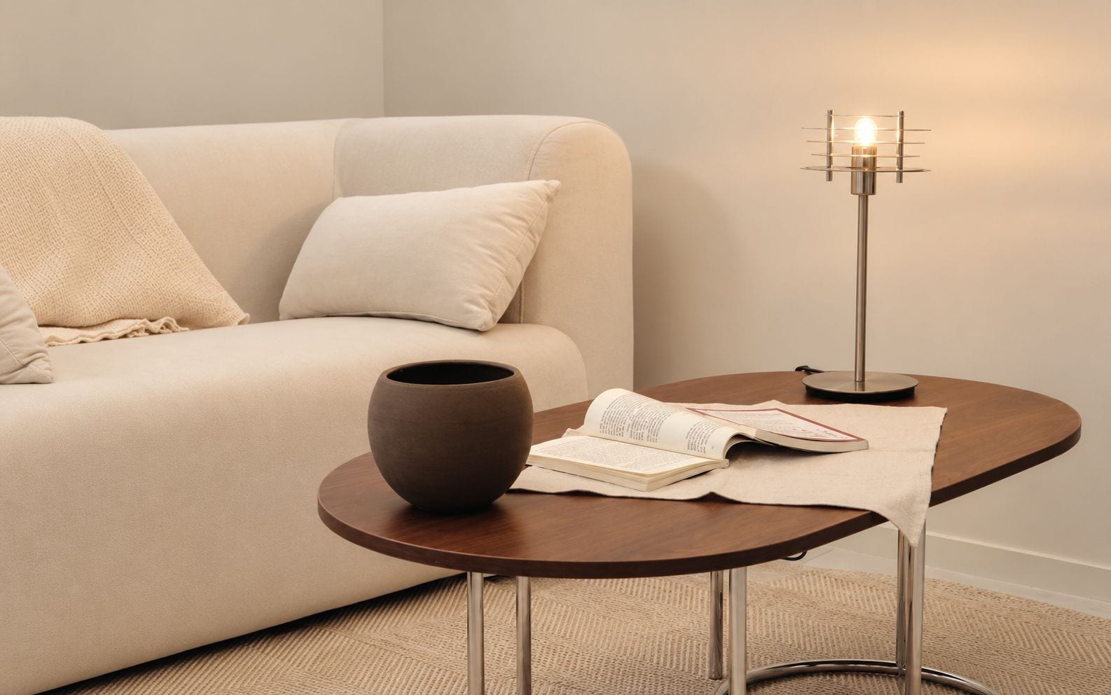 A modern living room with a beige sofa, a round wooden side table, a ceramic vase, an open book, and a floor lamp.