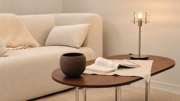 A modern living room with a beige sofa, a round wooden side table, a ceramic vase, an open book, and a floor lamp.