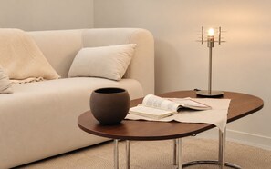 Una sala moderna con un sofá color beige, una mesita redonda de madera, un jarrón de cerámica, un libro abierto y una lámpara de pie.