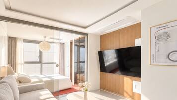 A modern hotel room with a large flat-screen TV, a sofa, a coffee table, and a view through a glass door.