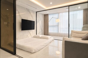 Una habitación de hotel moderna con una cama, un sofá, un televisor montado en la pared y un amplio ventanal con cortinas.