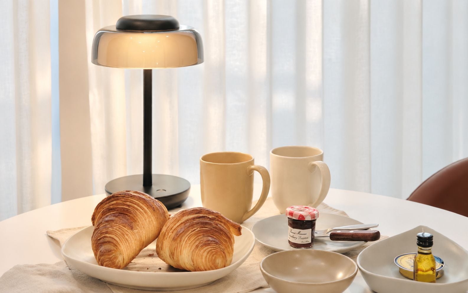 Een ontbijtsetting met croissants, koffiekopjes, een potje jam en een lamp.