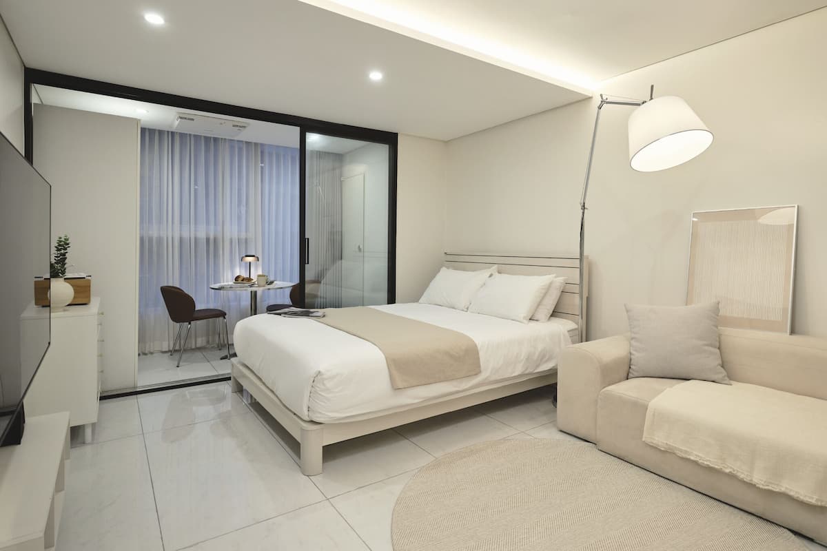 A modern hotel room with a bed, a sofa, a desk with a lamp, and a sliding glass door leading to a balcony.
