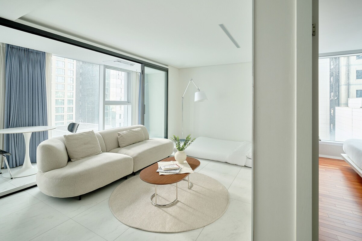 A modern living room with a sofa, a coffee table, a lamp, and a view of the city through large windows.