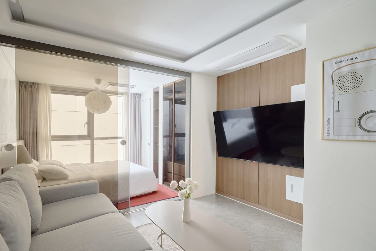 A modern hotel room with a large flat-screen TV, a sofa, a vase with flowers, and a sliding glass door leading to a balcony.