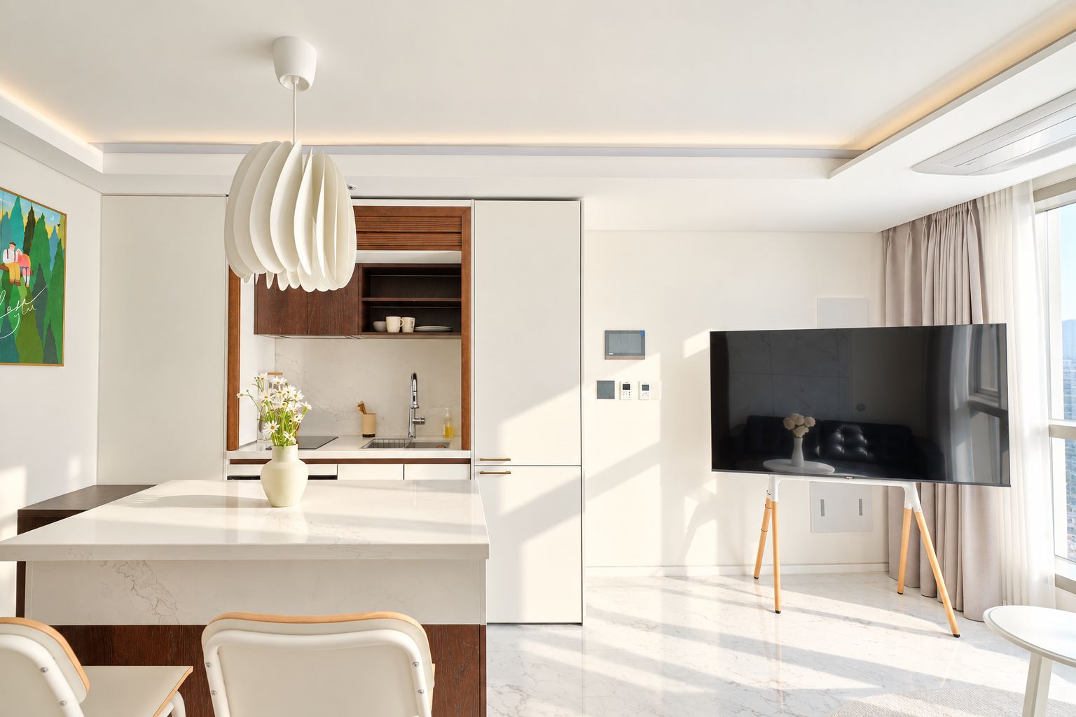 Cocina moderna con una isla central, zona de comedor con sillas, televisor de pantalla plana y un amplio ventanal.