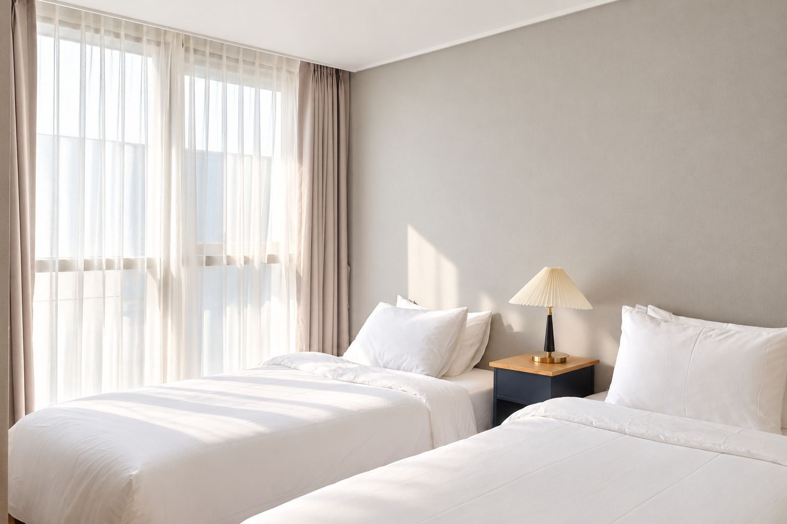A hotel room with two beds, a nightstand with a lamp, and sheer curtains on a window.