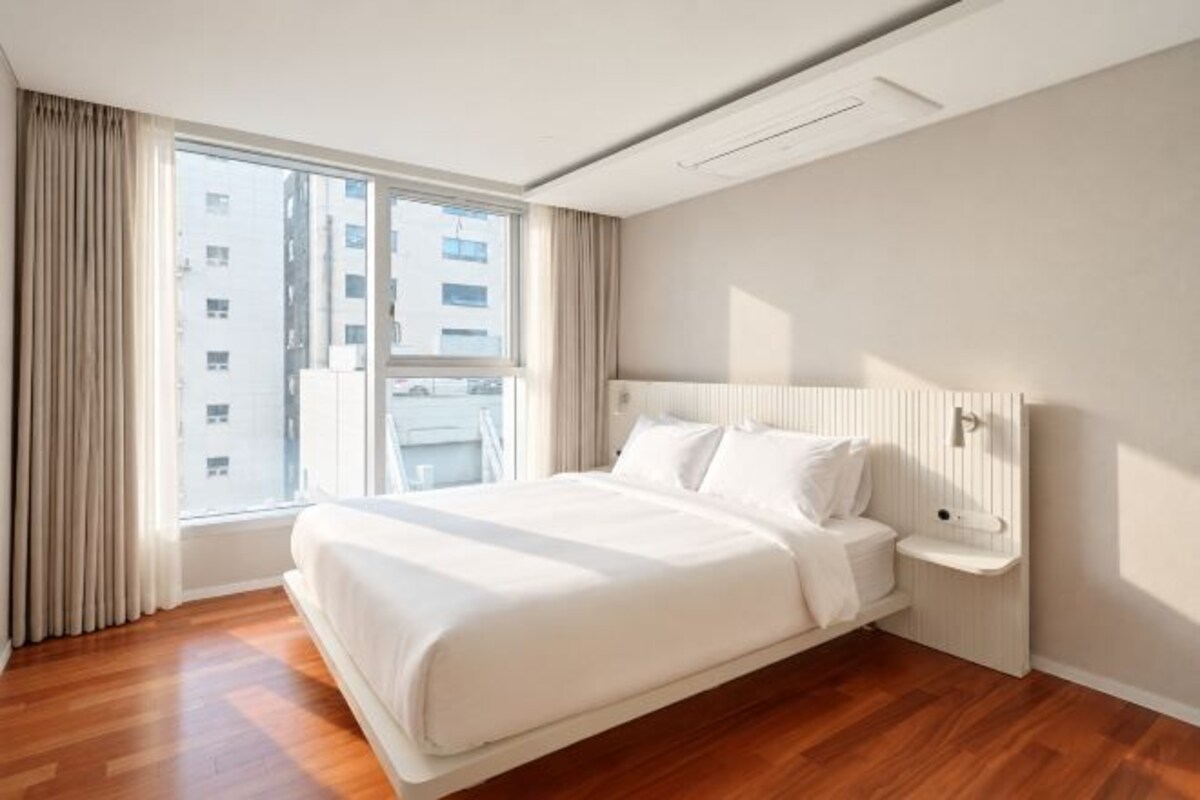 A hotel room with a large bed, a bedside table, a window with curtains, and a view of buildings outside.