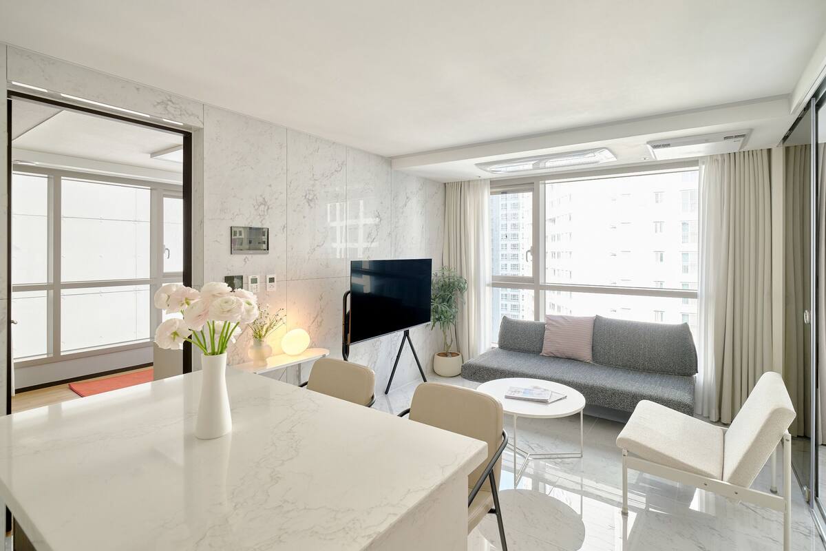 A modern hotel room with a marble-topped dining table, a sofa with cushions, a flat-screen TV, and a large window with curtains.