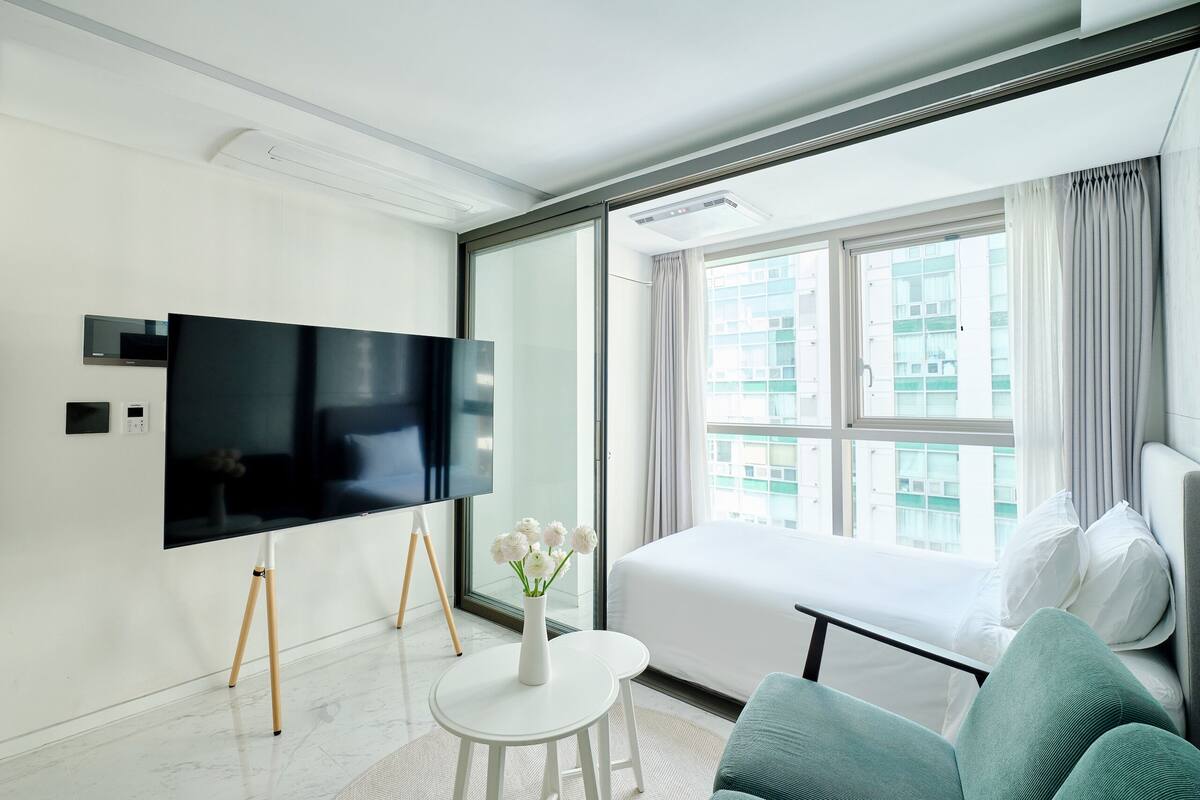 A modern hotel room with a large flat-screen TV, a bed, a sofa, and a small table with a vase of flowers.