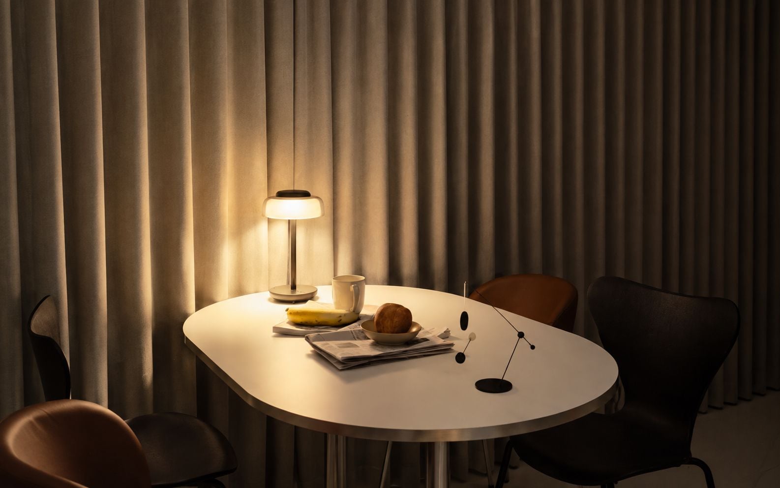 A round table with a lamp, a banana, a croissant, and a newspaper.