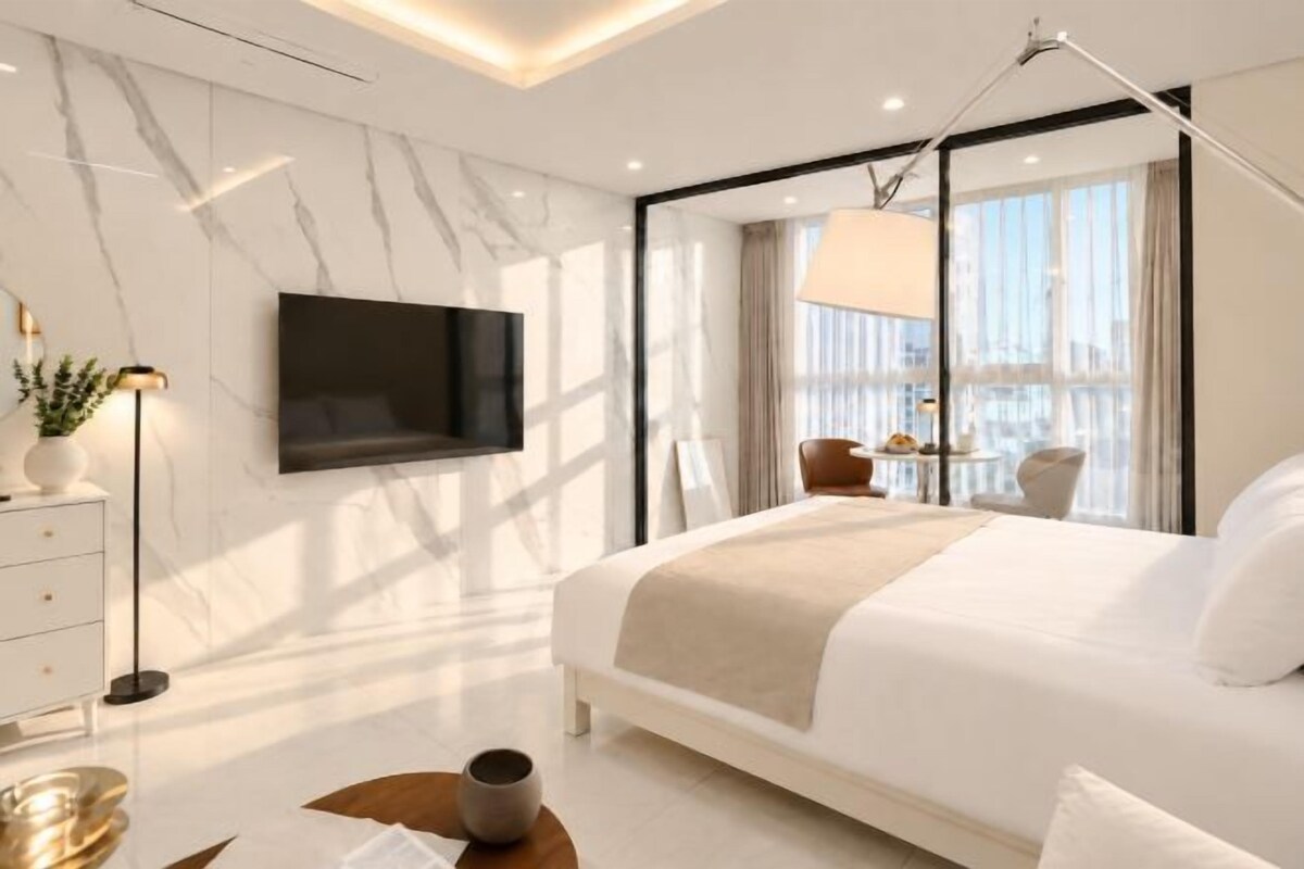 A modern hotel room with a large bed, a flat-screen TV mounted on the wall, a bedside table with a lamp, and a view of the city through large windows.