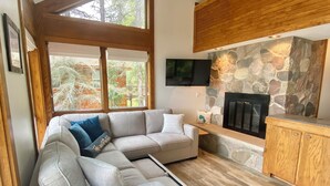 A modern living room with a sectional sofa, a stone fireplace, and large windows. - Beautiful Home w/ 2 fireplaces, Deck, and 75ft of Private Higgins Lake Frontage (Roscommon)