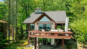 A modern two-story house with a large deck, surrounded by lush greenery. - Beautiful Home w/ 2 fireplaces, Deck, and 75ft of Private Higgins Lake Frontage (Roscommon)
