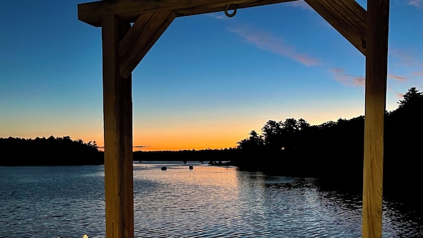 A wooden deck with a gazebo, a table, and a chair overlooking a calm body of water at sunset. - Large beautiful ocean front studio with private dock (Harpswell)