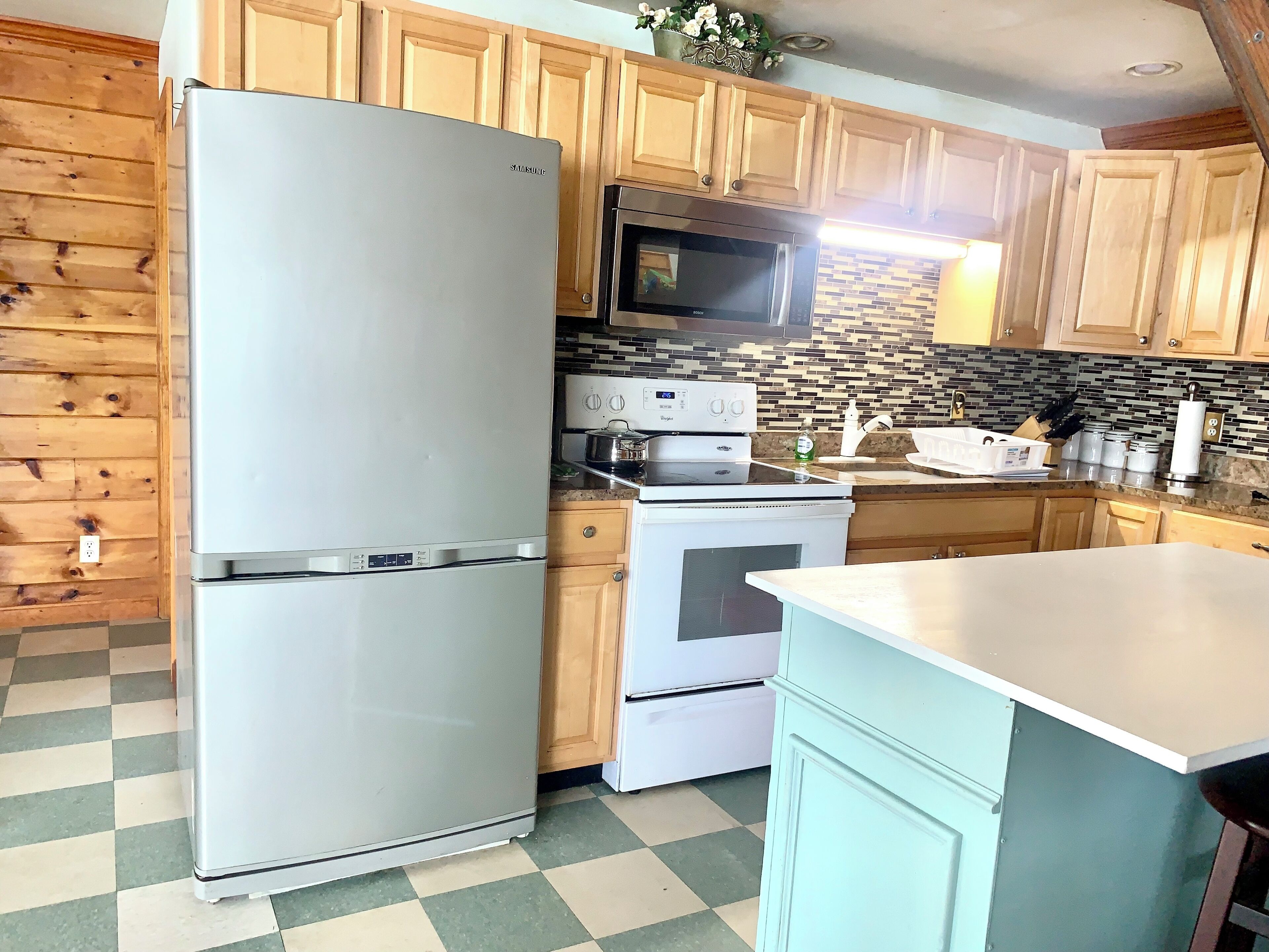 A kitchen with wooden cabinets, a stainless steel refrigerator, a microwave, an oven, and a checkered floor.
