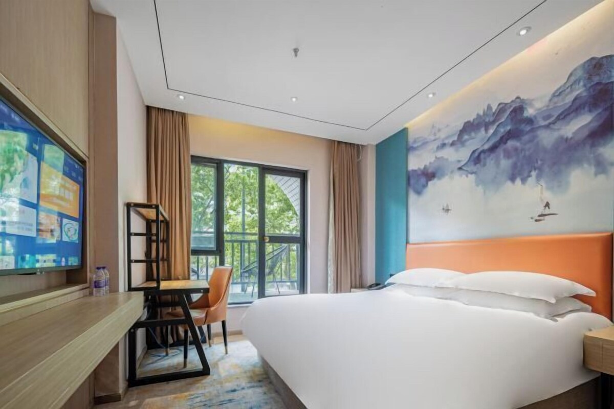 A hotel room with a large bed, a desk with a chair, a TV, and a window with a view of greenery.