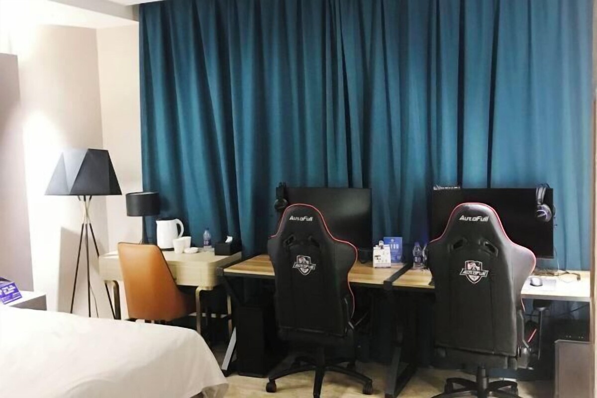 A hotel room with two gaming chairs, a desk, a laptop, a lamp, a bed, and a curtain.