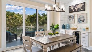 A spacious dining area with a large wooden table, comfortable chairs, and a view of a lush garden through large sliding glass doors. - Coastal Villas (Destin)