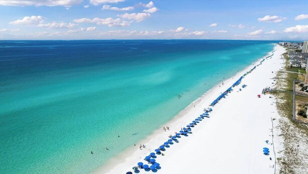 An aerial view of a beach with clear turquoise waters, a long stretch of white sand, and a row of blue umbrellas along the shore. - Coastal Villas (Destin)