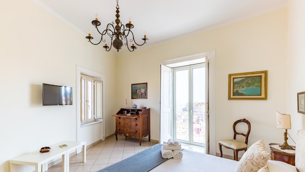 Habitación de hotel con cama, cómoda, televisor, banca, sillón y candelabro.