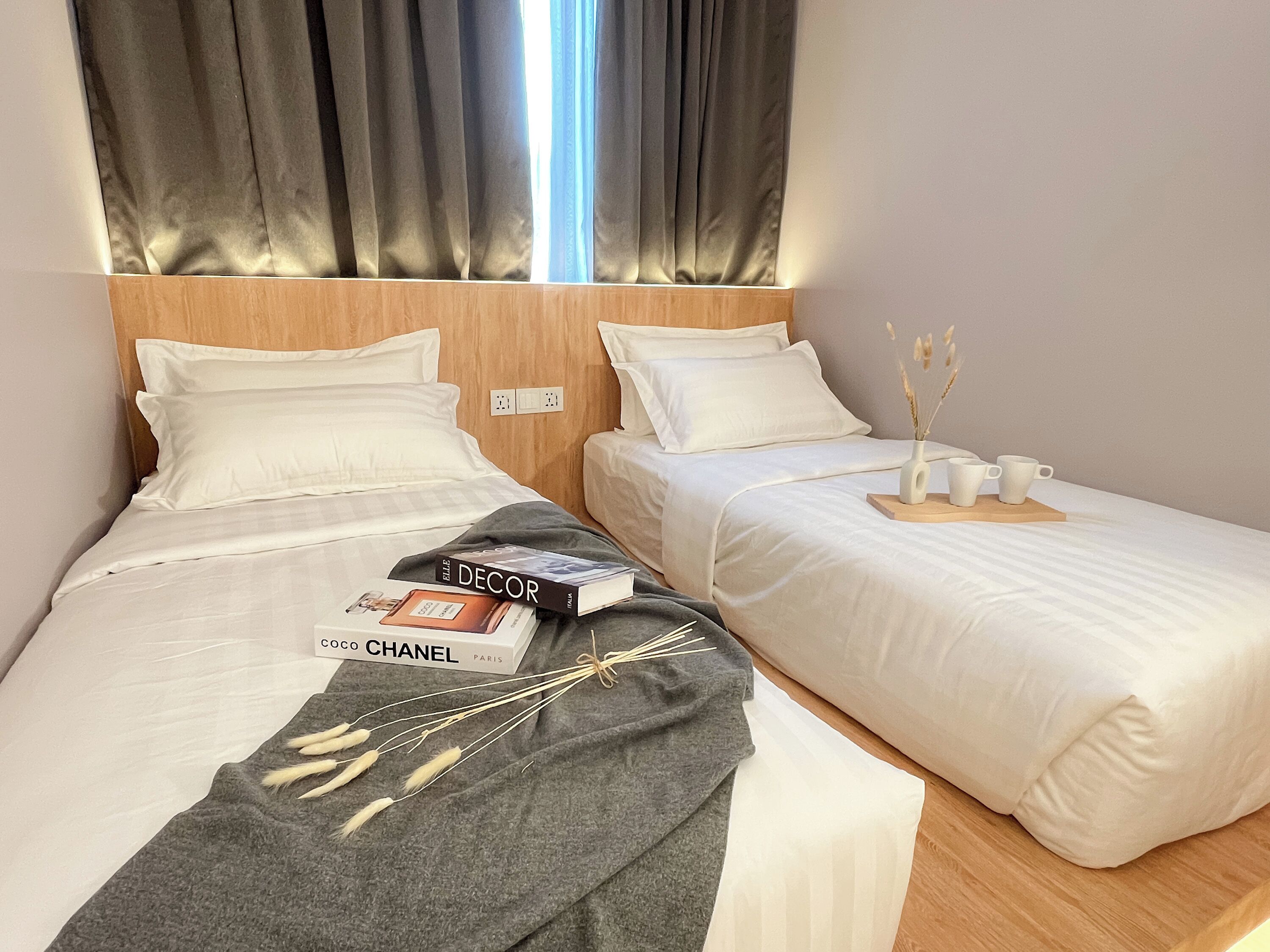 Quarto de hotel com duas camas, cada uma com roupa de cama e almofadas brancas, uma cabeceira de madeira e uma pequena mesa com um vaso e chávenas.