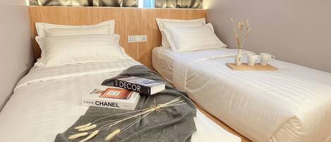A hotel room with two beds, each with white bedding and pillows, a wooden headboard, and a small table with a vase and cups.