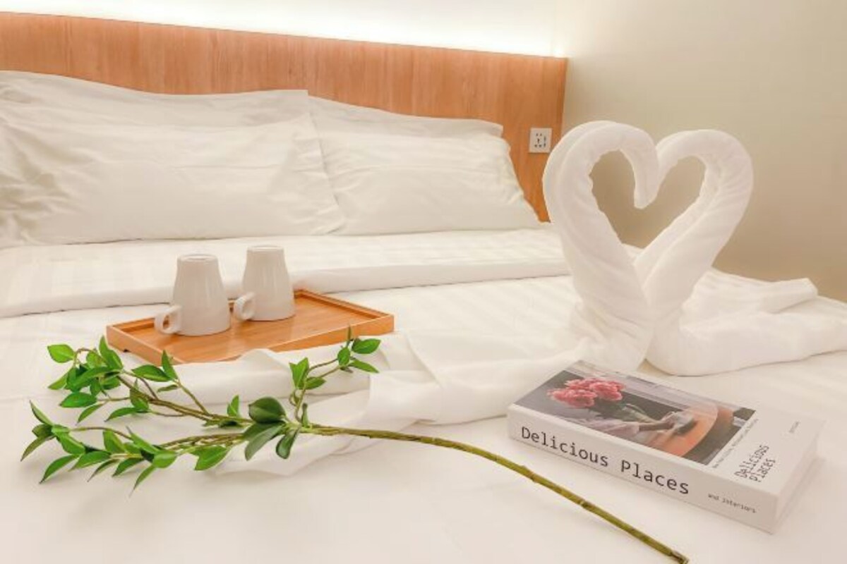 A neatly made bed with white linens, a tray with two white cups, a book titled 'Delicious Places', and a decorative swan made of fabric.