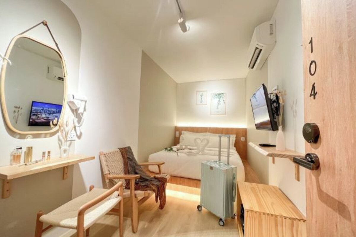 Een kleine hotelkamer met een bed, bureau, stoel, grote spiegel, televisie en koffer.