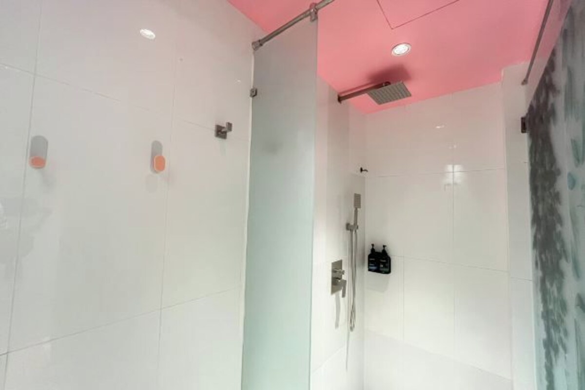 A modern bathroom with white tiles, a glass shower enclosure, and a pink ceiling.