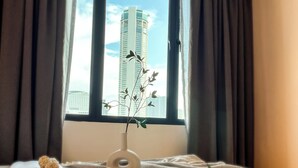 A bed with a vase of flowers, a view of a tall building, and dark curtains. - Sleep Box Penang (George Town)