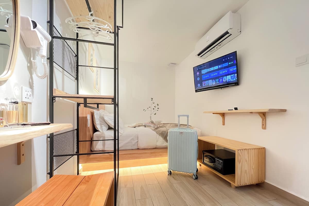 A compact hotel room with a bunk bed, a desk with a hairdryer, a TV mounted on the wall, a suitcase, and a small shelf.