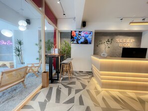 A modern reception area with a 'Sleep Box' sign, a reception desk, and a TV screen displaying a program. - Sleep Box Penang (George Town)