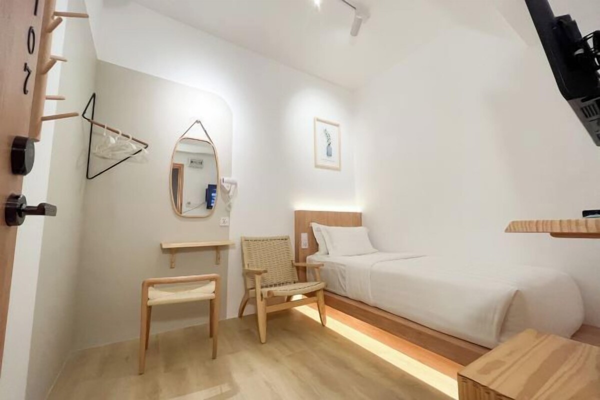 Habitación de hotel minimalista con cama, una silla, una mesita, un espejo y un cuadro en la pared.
