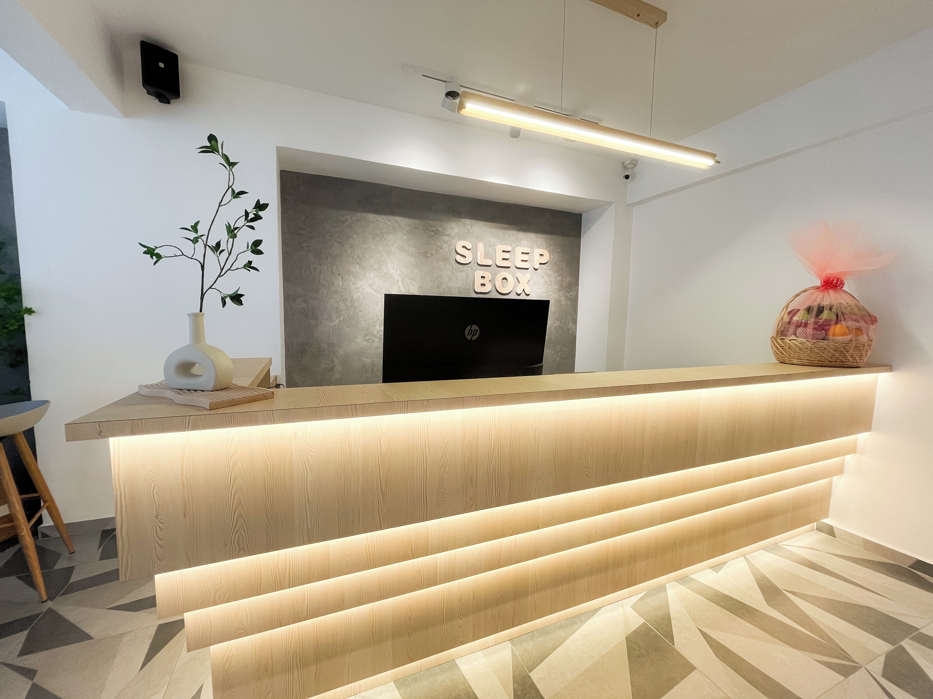 A reception desk with a 'SLEEP BOX' sign, a vase with a plant, and a basket of fruit.