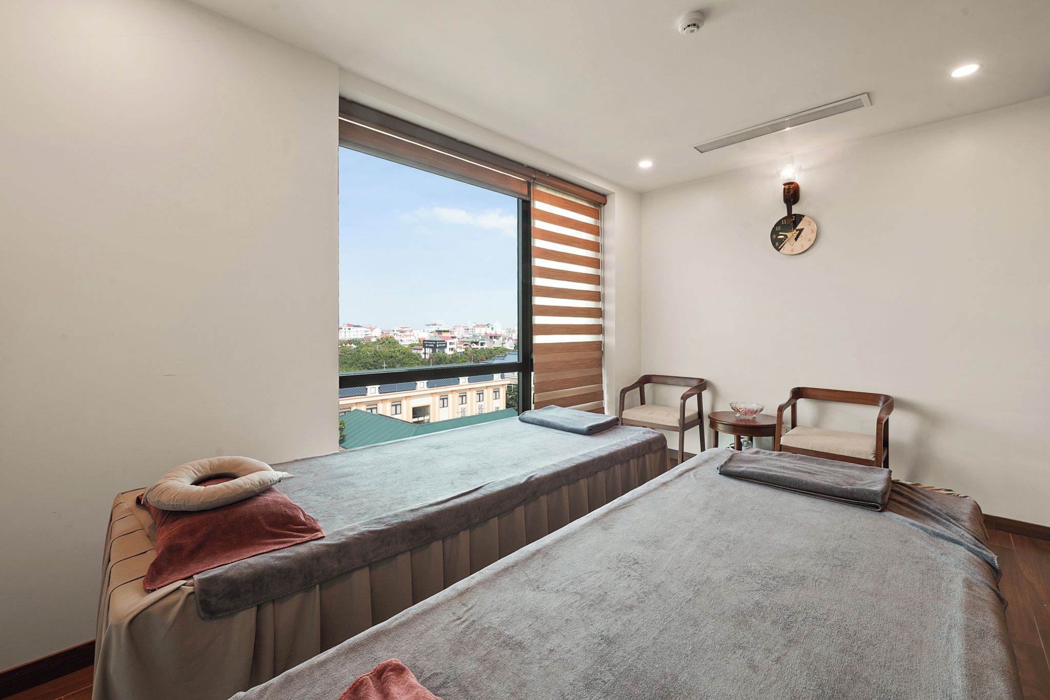 A room with two massage beds, a small table with a bowl, and a view of a cityscape through a large window.