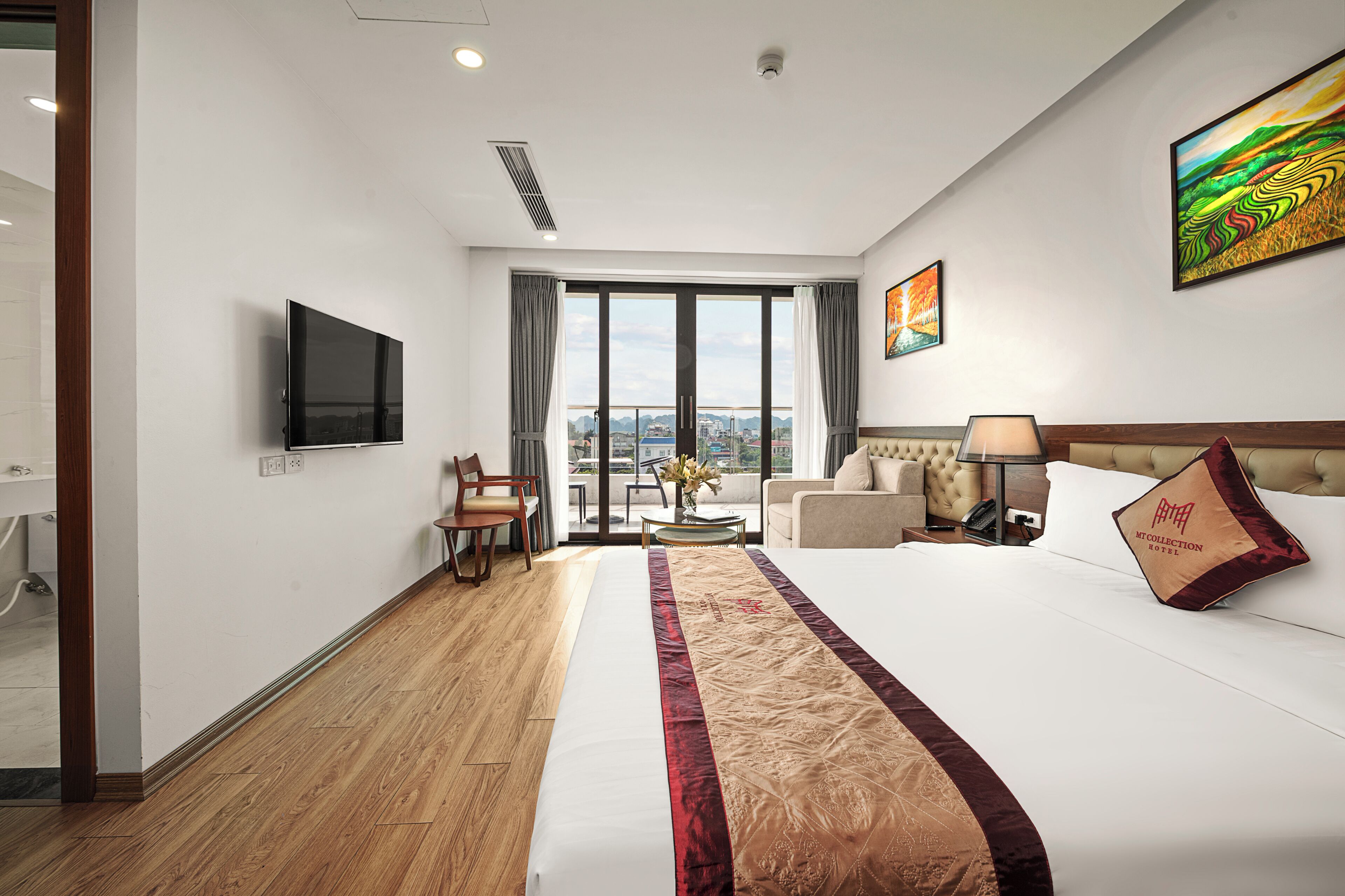 A hotel room with a large bed, a TV, a sofa, a chair, a table with a vase, and a view of the city through large windows.