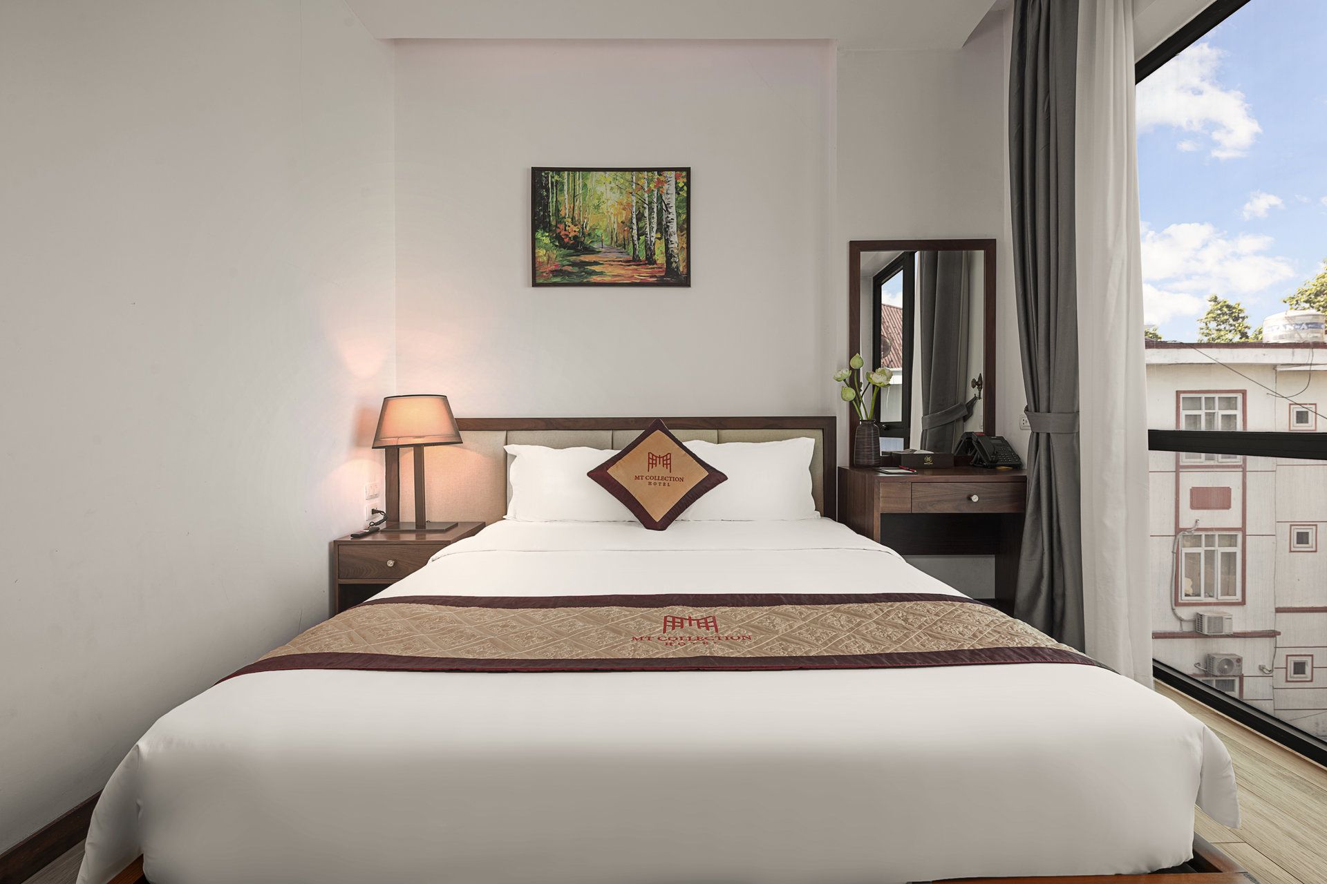 Habitación de hotel con cama, mesita de noche con lámpara, un cuadro en la pared, ventana con cortinas y vista a edificios.