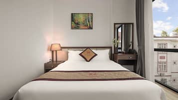 Habitación de hotel con cama, mesita de noche con lámpara, un cuadro en la pared, ventana con cortinas y vista a edificios.