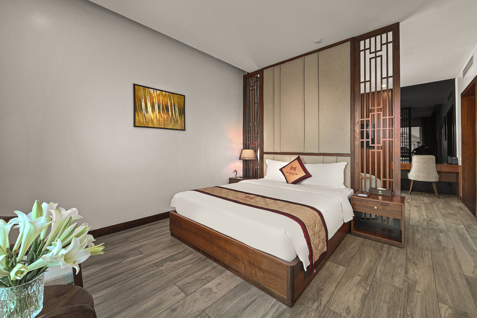 A hotel room with a large bed, a bedside table, a lamp, a decorative screen, and a vase with flowers.