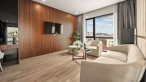 A modern living room with a wooden accent wall, a flat-screen TV, a sofa, a coffee table, and a view of the city. - MT Collection Hotel (Hoa Lu)