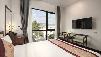 A hotel room with a large bed, a TV mounted on the wall, a desk with a telephone, and a view of the outside through a large window.