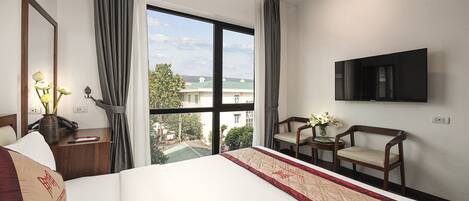 A hotel room with a large bed, a TV mounted on the wall, a desk with a telephone, and a view of the outside through a large window.