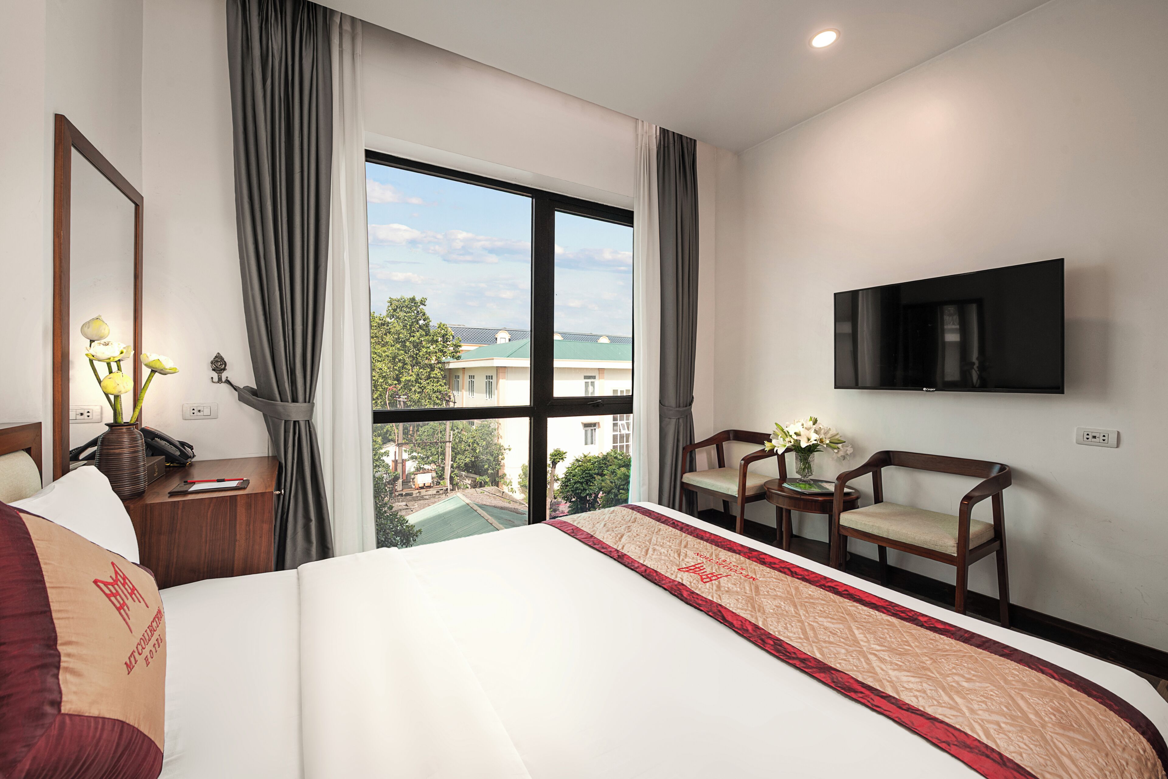 A hotel room with a large bed, a TV mounted on the wall, a desk with a telephone, and a view of the outside.