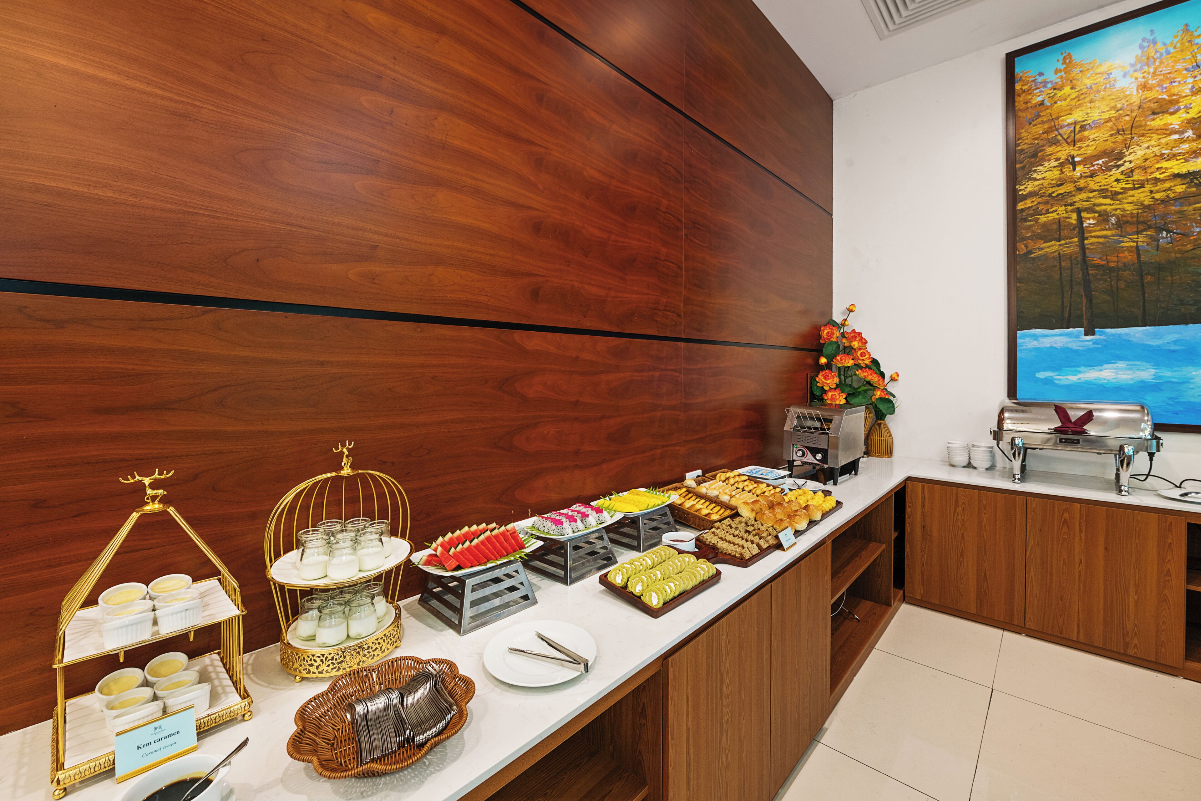 A buffet setup with various food items, a decorative golden stand, and a painting of a forest scene.