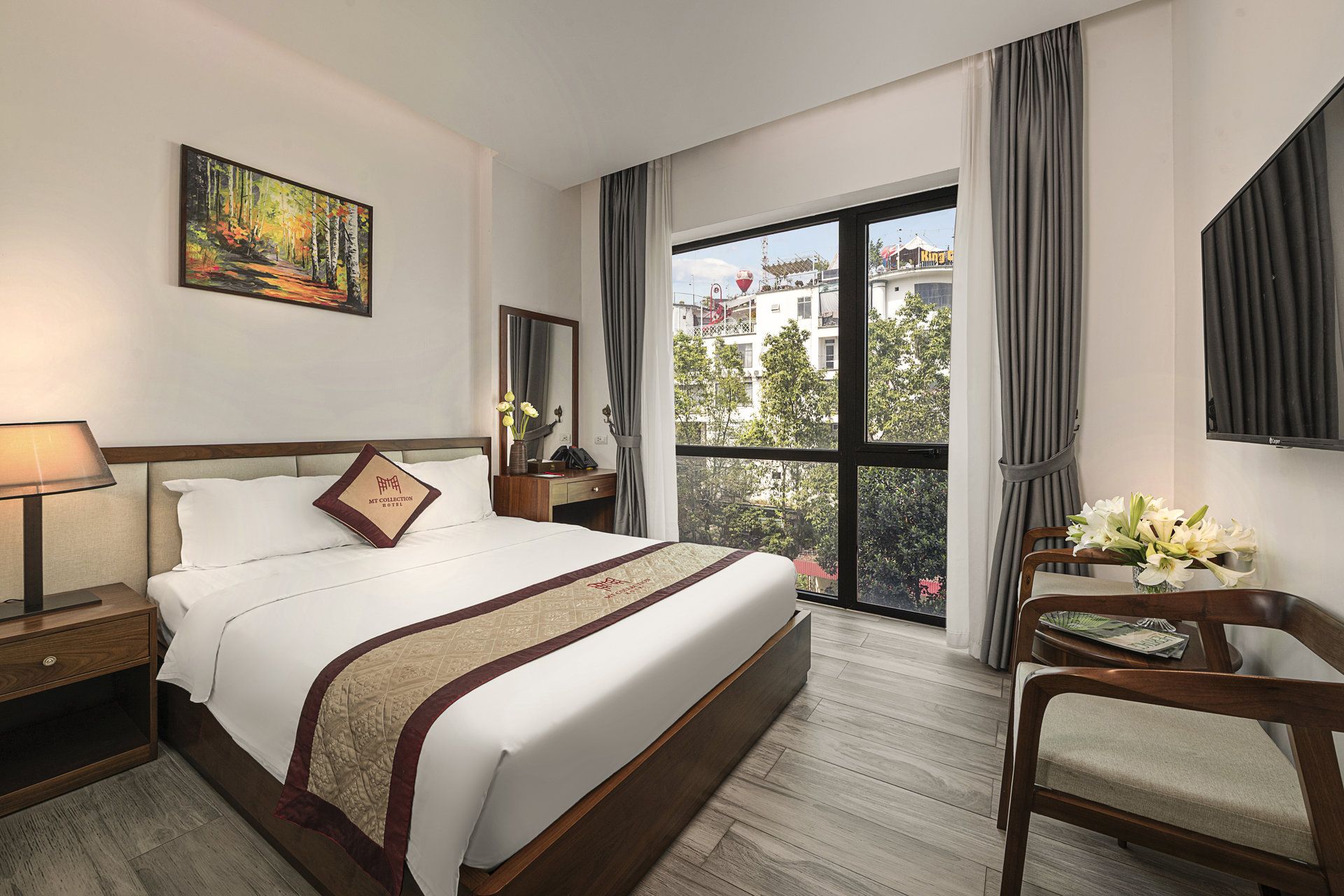 A hotel room with a large bed, a bedside table with a lamp, a chair, a desk with a vase of flowers, a large window with a view of buildings, and a painting on the wall.