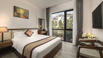 A hotel room with a large bed, a bedside table with a lamp, a chair, a desk with a vase of flowers, a large window with a view of buildings, and a painting on the wall.